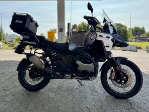 BMW R 1300 GS Adventure parkert på en bensinstasjon, vist i profil med stor toppboks, eikerhjul og den karakteristiske store tanken