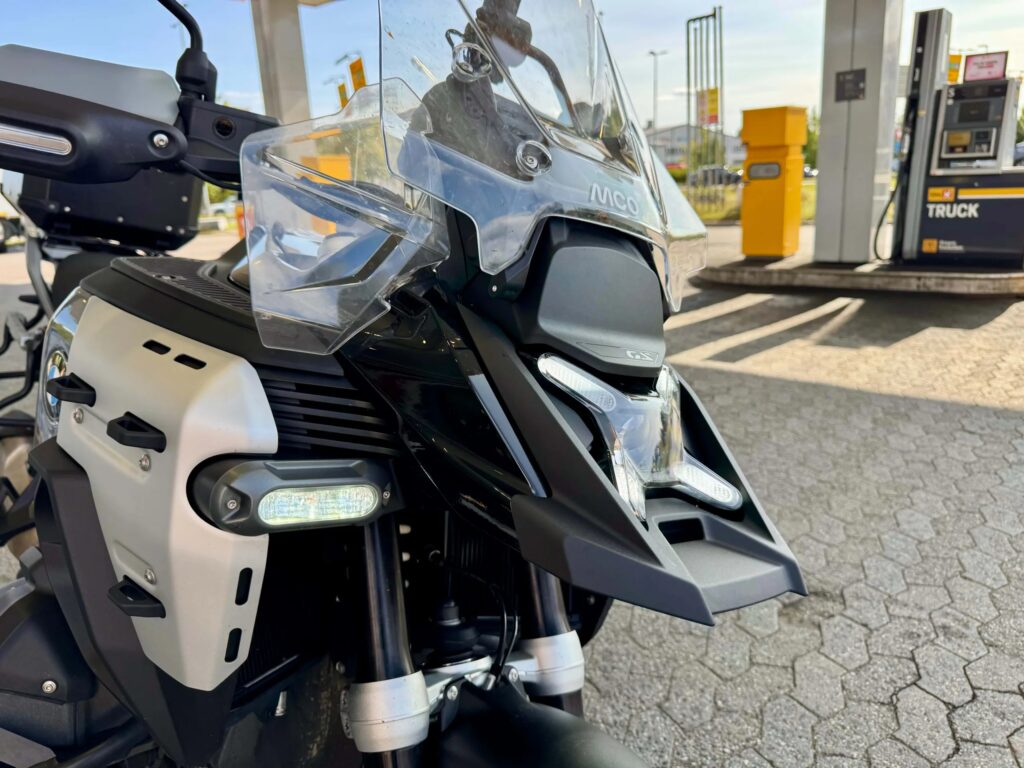 Nærbilde av fronten på BMW R 1300 GS Adventure med stor vindskjerm, ekstralys og festepunkter på tanken, fotografert ved en bensinstasjon