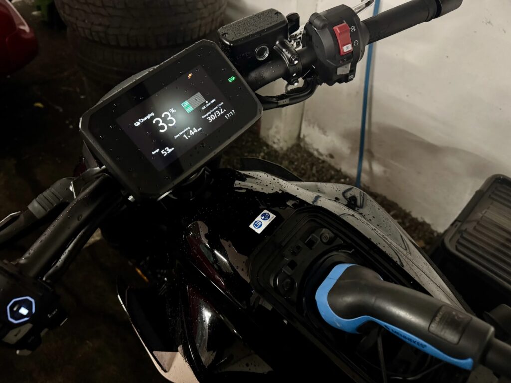 Nærbilde av Honda WN7s 5-tommers TFT-skjerm som viser 33 % ladestatus, 53 km rekkevidde og 1 t 44 min ladetid igjen, med en Type 2 hjemmelader blå CCS-ladekabel tilkoblet