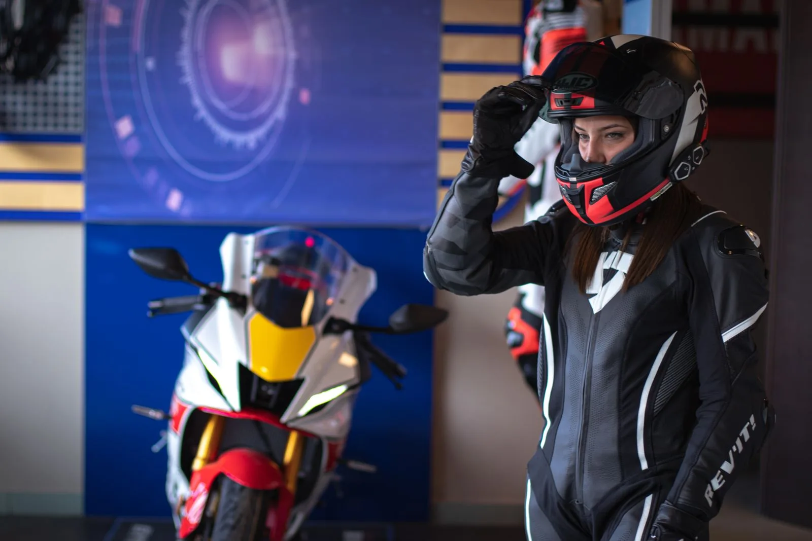 A woman motorcyclist in leather gear adjusts her helmet indoors.
