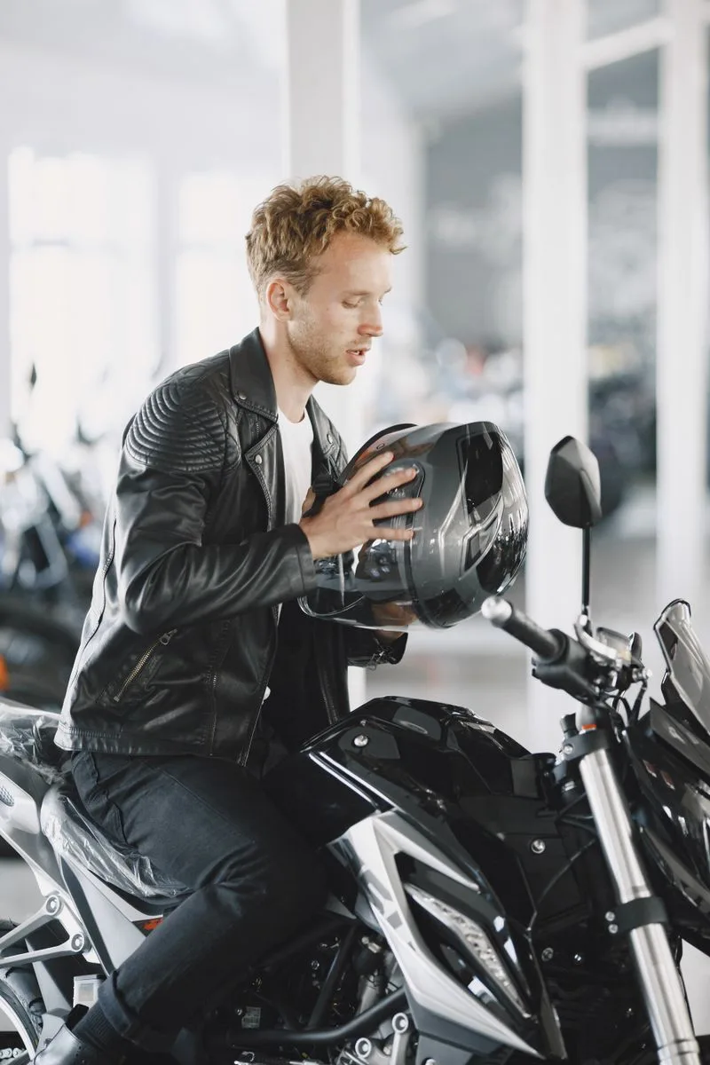 Adult man in leather jacket holding helmet, sitting on motorcycle indoors.